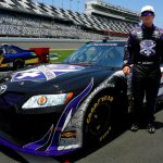 the infinite hero driver, wearing purple sunglasses, stands next to his car at Daytona Speedway